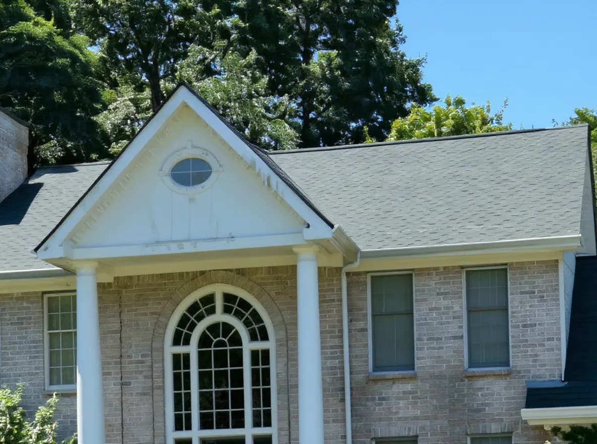 Closer view of the roof: a two-story brick house with a gray asphalt shingle roof and arched entryway, surrounded by a well-maintained lawn and mature trees