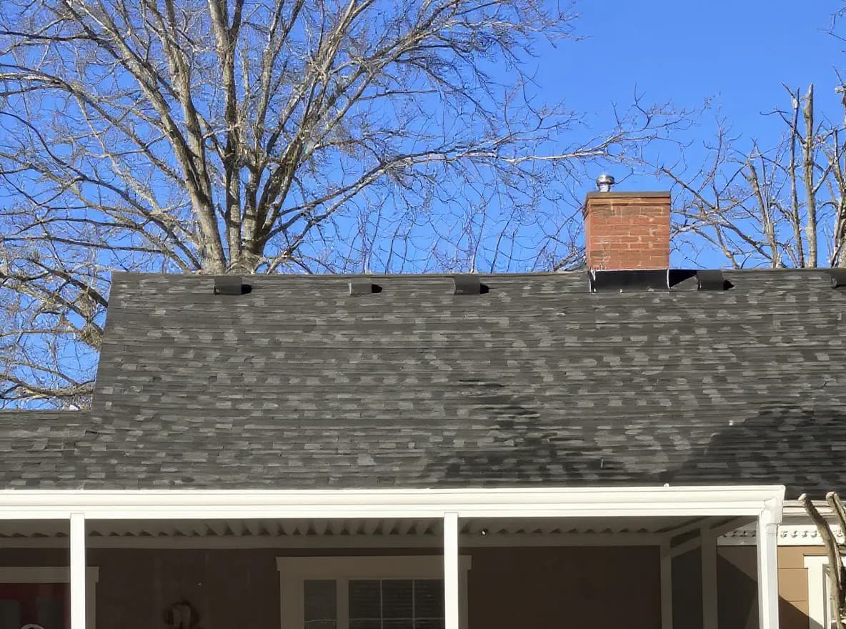 Closer view of the roof: rear exterior view of a single-story house with a screened-in porch, dark asphalt shingle roof, and brick chimney under a clear blue sky