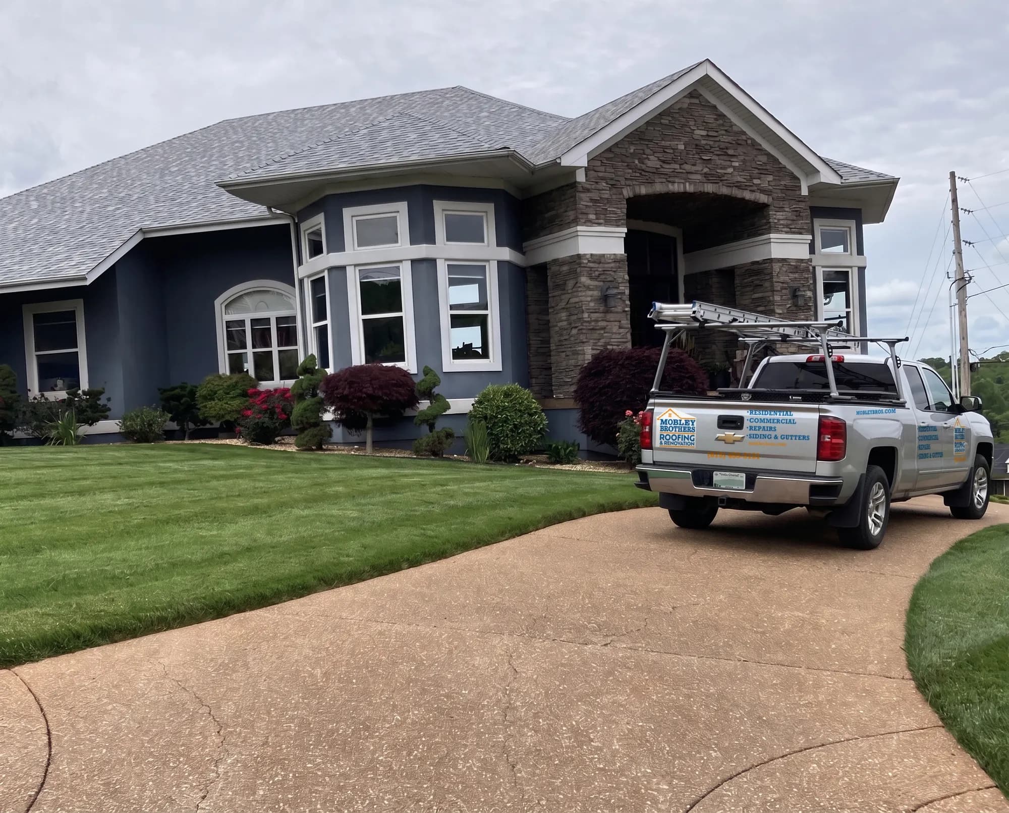 A Mobley Brothers Roofing branded truck with a ladder rack parked in the driveway of a stone and blue-sided home